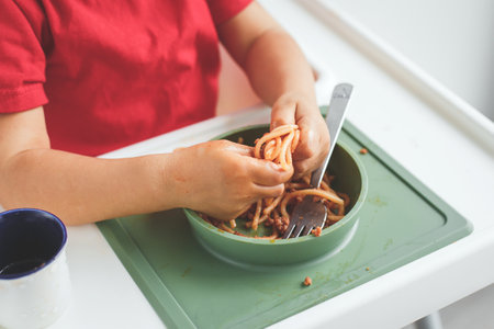 Child eating spaghetti with hands, making a mess on the high chair tray. The playful meal moment captures childhood, food, and family life conceptの写真素材