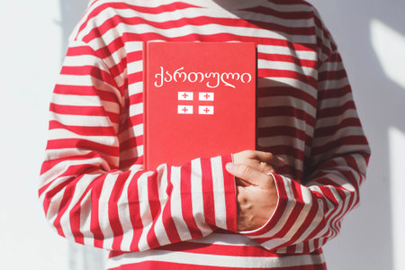 One girl, whose face is not visible in a striped red long sleeve, holds a red Georgian language textbook with Georgian flag and Georgian inscription in Georgian, foreign language learning concept.の写真素材