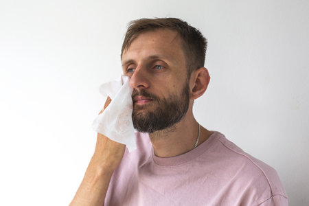 A bearded white young man in a pink T-shirt is using a facial cleansing wipe on his cheek, standing against a white wall in natural light. close up portraitの写真素材
