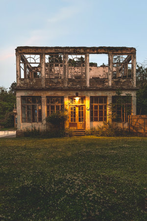 Abandoned two-story building with broken windows, glowing lantern above the entrance, surrounded by overgrown plants and silent twilight, verticalの写真素材