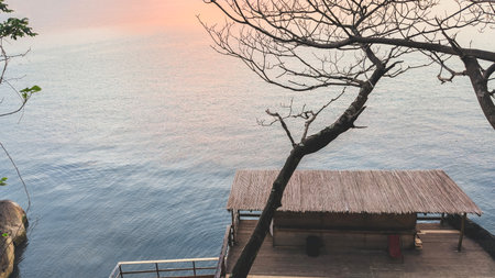 Wooden hut with thatched roof on waterfront terrace, surrounded by calm sea and tree branches at sunset, peaceful tropical retreat atmosphere, concept of travel and relaxationの写真素材
