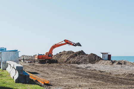 An orange excavator makes a mound of sand on the seashore. Protection of the beach area from flooding. Industrial construction equipment and machinery. repair of the embankment after the storm.の写真素材