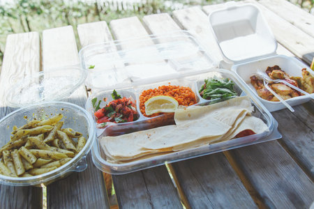 A variety of Turkish takeout food served outdoors on a wooden picnic table, including bulgur with lemon, salad, lavash, grilled chicken wings, pasta with pesto, and fresh herbs.の写真素材