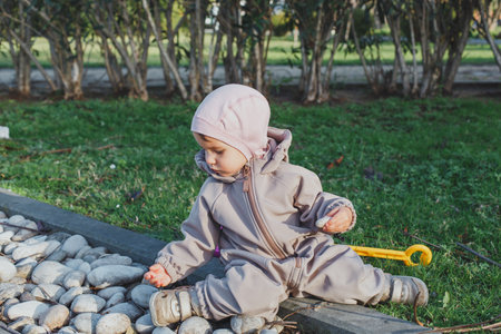 A small child girl 1-2 year in a warm jumpsuit and hat sits on the ground in a park, playing with...
