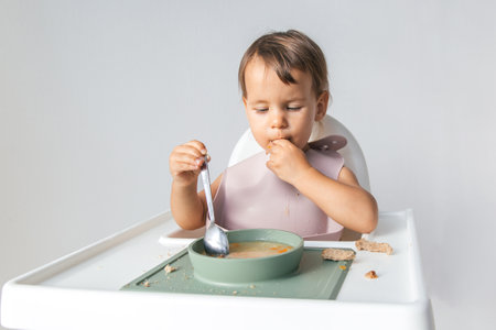 one baby 1 year and 3 months old sits in a high chair on a white background and eats soup with bread.の写真素材