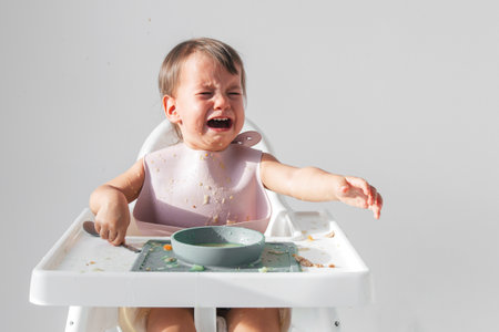 Crying toddler sitting in a high chair, covered in food, reaching out with one hand while holding a spoon in the other, expressing frustration during mealtime in a bright home settingの写真素材