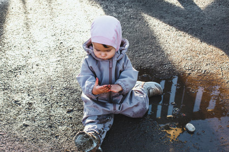 Toddler in a waterproof suit and hat sitting on wet ground, curiously examining muddy hands after playing in a puddle, surrounded by reflections and soft sunlightの写真素材