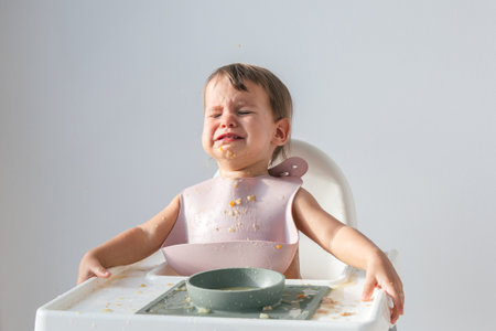 cute crying baby sitting eating lunch in high chair at home on white background, hysterics and tears...