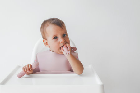 close-up portrait of a cute baby girl infanta 8 months old, sitting on a highchair in a baby apron with a fork and spoon, the concept of baby food and baby food.の写真素材