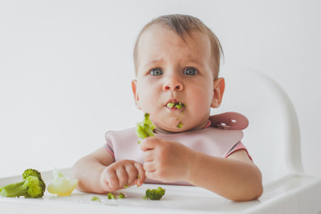 Child small and cute close-up portrait sits in a highchair on a white background and eats broccoli complementary foodsの写真素材