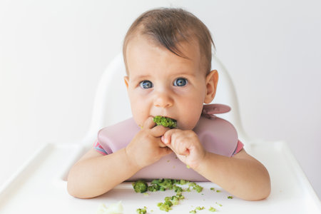 baby little girl 8 months old sits in a high chair and eats complementary foods green broccoli, close-up portrait looks at the camera. baby food conceptの写真素材