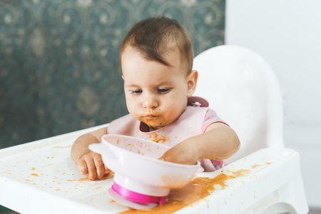 baby 9 months old sits in a highchair and eats complementary foods vegetable puree soup spilled from a plate, the concept of baby food.の写真素材