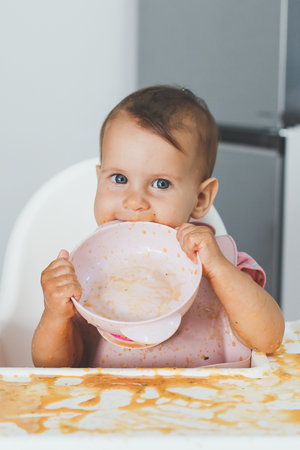 funny cheerful dirty baby sits in a highchair in the kitchen and eats complementary foods, holds an...