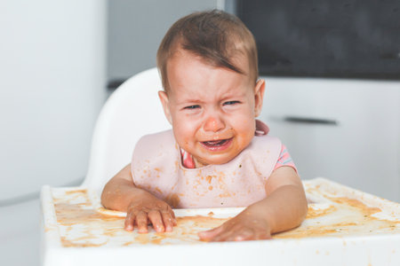 small cute baby 9 months old sits in the kitchen in a high chair and cries, grimy and dirty, spilled food.の写真素材