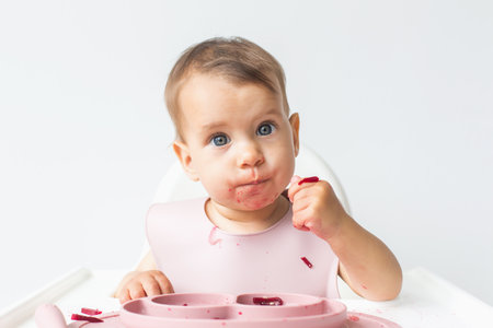 close-up portrait of a cute little girl 9 months old sitting in a highchair in the kitchen, looking at the camera, eating complementary foods.の写真素材
