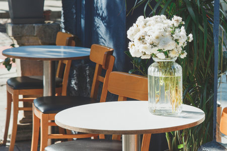 Cozy outdoor caf with round tables and wooden chairs, featuring a glass vase with fresh white flowers, creating a warm and inviting atmosphere in natural sunlightの写真素材