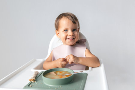 one baby 1 year and 3 months old sits in a high chair on a white background and eats soup with bread, smiles and looks at the camera, complementary feeding concept.の写真素材