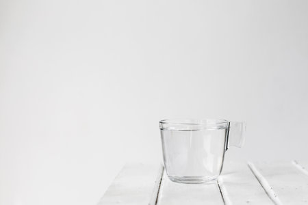 A clear glass cup filled with water sits on a white wooden surface against a minimalist white background. The scene emphasizes purity, simplicity, and clarity. copyspaceの写真素材