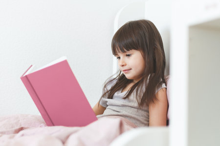 one todder girl 5 years old brunette with long hair sits in bed and reads a book.の写真素材