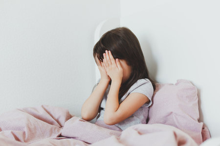 brunette toddler girl sitting in bed, covering her face with her hands, crying, frustrated.の写真素材