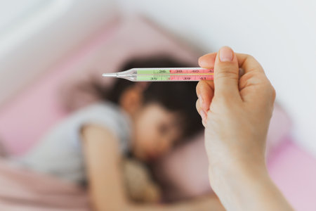 mom holds a thermometer with a high temperature of 39 near the bed of a sleeping child, the child is out of focus, the concept of childhood illnessの写真素材