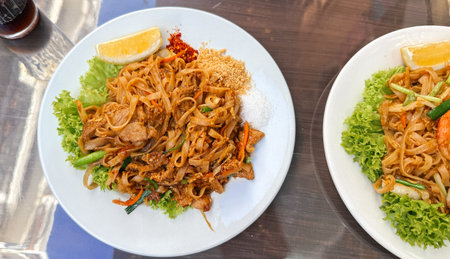 A plate of Thai Pad Thai noodles with chicken, garnished with lettuce, peanuts, chili flakes, sugar,...