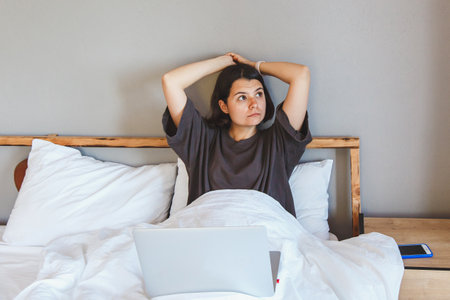 Young woman in casual clothes sitting in bed with a laptop, stretching and looking tired, suggesting...