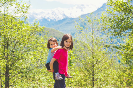Two young girls sisters enjoy a piggyback ride in a lush green forest with snow-capped mountains in the background, radiating joy and sibling bonding in nature.の写真素材
