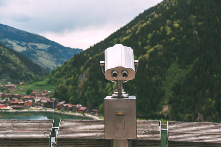 A coin-operated binocular viewer overlooks a scenic mountain valley and lake, offering panoramic views of a picturesque village surrounded by lush green hills.の写真素材