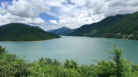 A wide turquoise lake is nestled between lush green mountains under a dramatic sky with thick...
