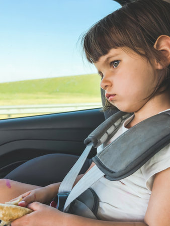 little girl sits quietly in a car seat, looking out the window with a pensive expression, holding a piece of bread in her hands during a road trip, travel conceptの写真素材