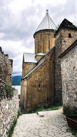 Ancient stone church with conical dome and narrow arched windows, surrounded by old fortress walls and cobblestone path, set against cloudy sky and forested mountains, travel conceptの写真素材
