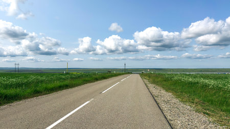 Straight empty road stretching into the horizon through green fields under a blue sky with scattered clouds, evoking freedom and peaceful solitude, travel conceptの写真素材