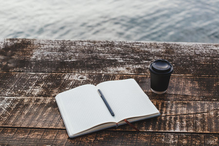A grid notebook with a pen lies open on a rustic wooden table by the water, next to a black takeaway...