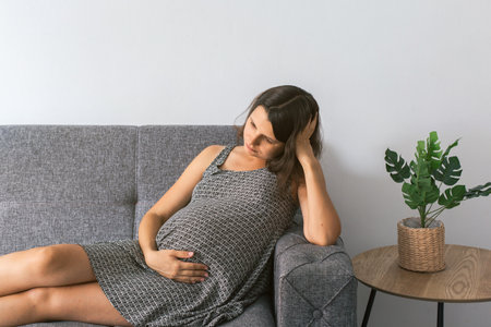 Pregnant woman lying on a gray couch with hand on belly and thoughtful expression, wearing patterned dress, next to a side table with potted plant, indoor scene, maternity conceptの写真素材