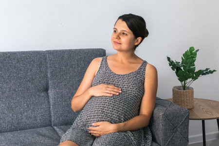 Pregnant woman sitting on gray couch with hands on belly and calm smile, looking away in peaceful mood, dressed in patterned sleeveless dress, indoor setting with potted plant, maternity conceptの写真素材