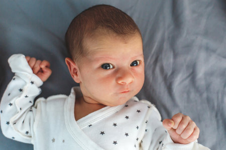 Newborn baby with raised fists lying on gray blanket, wearing white star-printed clothes, looking...