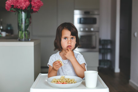 Little girl eating macaroni with fingers and looking forward with curious expression, sitting at the table in cozy kitchen, concept of childhoodの写真素材