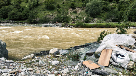 Piles of plastic, wood, and broken glass litter the rocky bank of a fast-flowing muddy river, surrounded by green forested hills, illustrating environmental neglect, pollution conceptの写真素材
