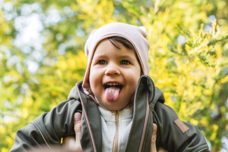Happy child sticking out tongue while being lifted in the air, wearing warm clothes and a beanie, playful moment in nature, surrounded by blooming mimosa flowers, joyful springtime sceneの写真素材