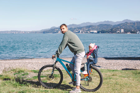 A father riding a bicycle with his young child seated in a safety seat on the back. They are enjoying a scenic outdoor ride near a beautiful lake with mountains in the background on a sunny dayの写真素材