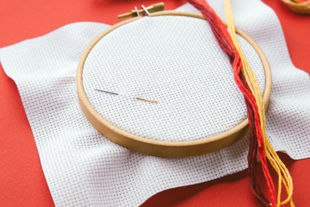 Embroidery hoop with white canvas, two needles and colorful threads on red background, flat lay of cross-stitching tools, concept of handmade craft and creativity conceptの写真素材