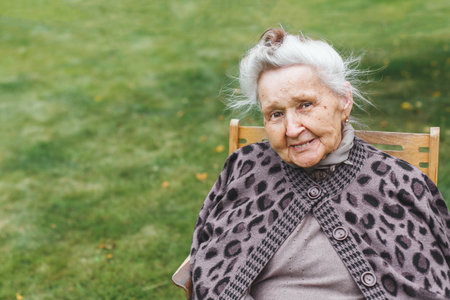 Smiling elderly woman 80-90 years with gray hair sitting on a wooden chair outdoors, wearing warm patterned sweater, concept of aging and positive lifestyle, copyspaceの写真素材