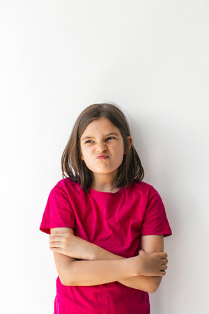 Angry capricious dissatisfied little child 8-9 years girl with arms crossed and a grumpy expression standing against white wall, concept of childhood emotions and mood, vertical portraitの写真素材