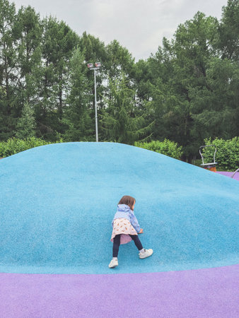 Little girl climbing a bright blue rubber hill at colorful modern playground surrounded by green trees, concept of childhood adventure and active outdoor playの写真素材