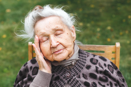 Elderly woman with closed eyes touching her head, expressing fatigue or discomfort, sitting outdoors, concept of aging and health awarenessの写真素材