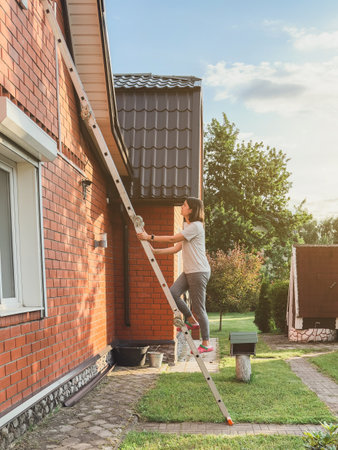 Woman climbing a ladder leaning on a red brick house during sunny day in garden, concept of home maintenance and independent lifestyleの写真素材