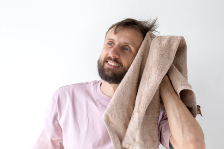 Smiling man wiping hair with beige towel after shower, wearing pink t-shirt, concept of self-care, freshness and morning routineの写真素材