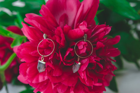 Golden hoop earrings with quartz crystal charms displayed on vibrant red peony flower, concept of beauty, fashion and natural eleganceの写真素材