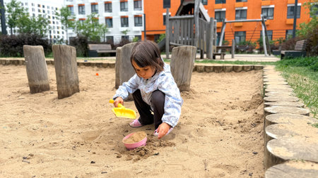 Little girl playing with sand in a modern urban playground, wearing a light jacket and using toy shovel and bucket, concept of childhood and outdoor activityの写真素材
